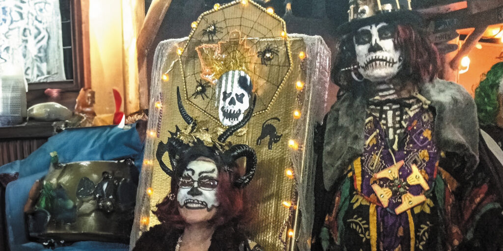 Patricia Perez has black-and-white skull makeup on her face and a black horned headpiece covering most of her chin-length hair. She sits on what appears to be a spooky throne with a backrest shaped like a headstone and decorated with a skull, spiders, and string lights. Her husband Dave stands beside her with black-and-white skull makeup on his face, wearing a top hat, colorful purple and gold tunic, and black cape.