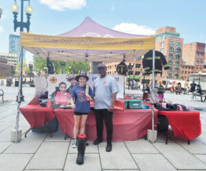 Breylyn, a young woman, stands in front of an outdoor Muscular Dystrophy Association display table wearing a fire fighter’s hat and holding the end of a fire hose. Beside her is a man wearing a International Association of Fire Fighters T-shirt.