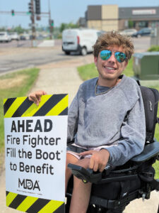 Luke, a young man, sits outdoors in a power wheelchair wearing mirrored aviator glasses and holding a sign that says “AHEAD Fire Fighter Fill the Boot to Benefit MDA.”