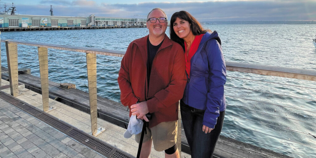 A.J. Bardzilowski lives in California with his wife, Marcey stand on a boardwalk beside a body of water. A.J. holds a cane for support.