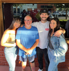 A man with white hair holding a can is surrounded by three young adults with their arms around each other.