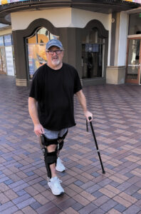 A.J. walks in an outdoor shopping area wearing black leg braces that extend from the upper thigh to the ankles and holding a black cane.