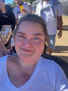 Closeup of Dana Carpenter, a woman with light skin, freckles, and light brown hair, at an outdoor festival. A line of stick-on rhinestones runs from her right cheek to above her left eyebrow. 