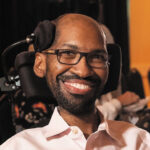 Closeup of Ira Walker, a man with brown skin and short black hair and beard wearing black-rimmed glasses. He sits in a wheelchair with a headrest.