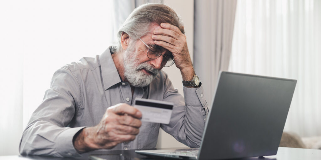 Upset senior elderly man holding credit card by laptop having trouble worry finance safety data or online payment security. Bank client concerned about problem with credit card, financial fraud threat