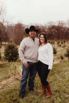 A man in a black cowboy hat and tan shirt and jeans smiles with his arm around a woman with brown hair in a white sweater and jeans 