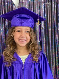 A young girl with curly hair smiles wearing a cap and gown for pre-K graduation