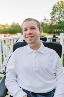 A young man in a white button down shirt smiles while seated in a power wheelchair