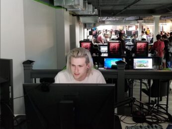 A young man with long blonde hair sits in front of a computer screen 