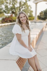 Abigail Phillips, a woman with light skin and long blond hair, poses sitting on the brick wall of an outdoor water fountain. She is wearing gold hoop earrings and a white knee-length dress. 