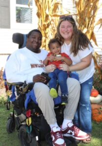 Justin Lopez, a man with dark brown skin and short black hair, sits in a wheelchair with is son, a toddler with medium brown skin and short curly brown hair, in his lap and his wife, Lexi, a woman with light skin and long brown hair, standing beside him. The family is outdoors with a corn stalk and stacked pumpkins in the background.