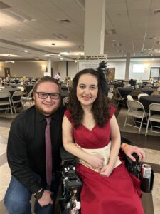 Olivia Holler, a woman with light skin and long, curly dark brown hair, sits in a black power wheelchair wearing a long red dress. Her boyfriend, Jacob, a man with light skin and short light brown hair and beard, kneels to the left of the wheelchair, wearing black glasses, black button-down shirt, and purple tie.