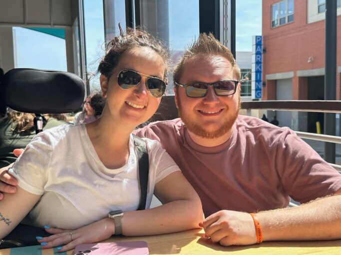 A couple wearing sunglasses sits at a restaurant table beside an open window on a sunny day.