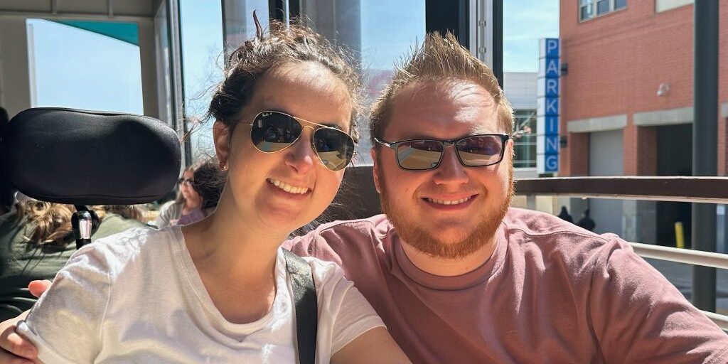 A couple wearing sunglasses sits at a restaurant table beside an open window on a sunny day.