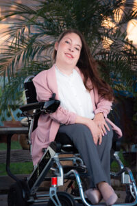 Briana Mills, a woman with light skin and long brown hair is sitting in a power wheelchair wearing a professional suit of pink jacket and gray slacks in front of a background of palm fronds.