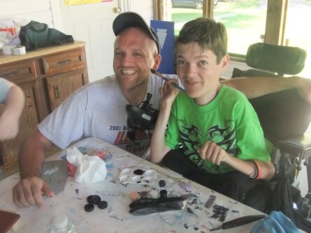 A man in a baseball hat kneels next to a young boy in a wheelchair at a craft table