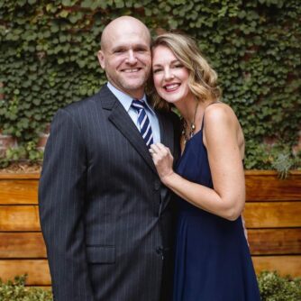 A man in a suit smiles as woman in a black dress leans against him in half an embrace