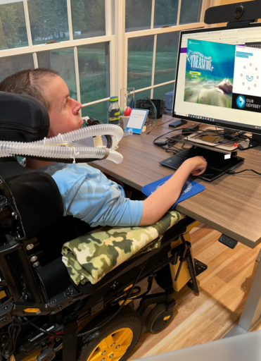 A young man in a power wheelchair uses an accessible video game controller to play games on a monitor screen