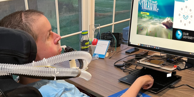 A young man in a power wheelchair uses an accessible video game controller to play games on a monitor screen