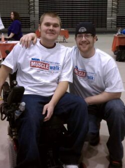 Two young men, one in a wheelchair and one squatting next to him, wear matching shirts at an MDA Muscle Walk