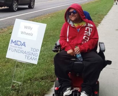 A man wearing a red sweatshirt and sitting in a wheelchair smiles next to a sign that says Willy Wheelz at an MDA Muscle Walk