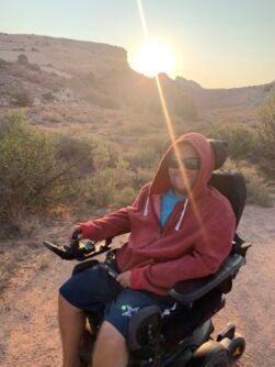 A young man in a wheelchair smiles wearing a red sweatshirt as the sun shines from between mountains behind him 