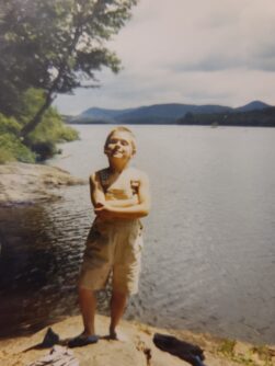 A young boy stands with his arms crossed in front of a lake