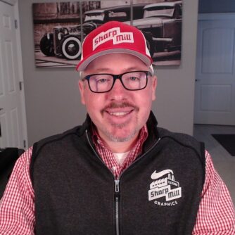 A headshot of a man wearing a branded Sharp Mill baseball hat and vest, smiling with glasses and goat-too facial har