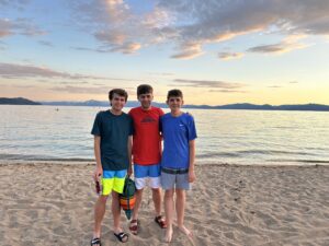 Three young adult men stand on a sandy beach in front of the ocean at sunset.