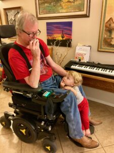 Jeffrey Gibler sits in a wheelchair playing the harmonica while his young grandson stands at his feet while watching and listening.