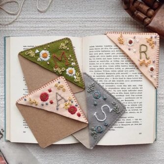 Embroidered bookmarks with flowers and one letter initial for the corner of a book page rest on an open book