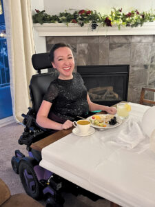 Amber Bosselman sits in a wheelchair at a dining table with a plate of food in front of her.