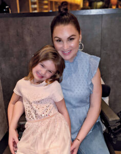 Cassidy Nilles sits with her 6-year-old daughter in her lap, both smiling at the camera.