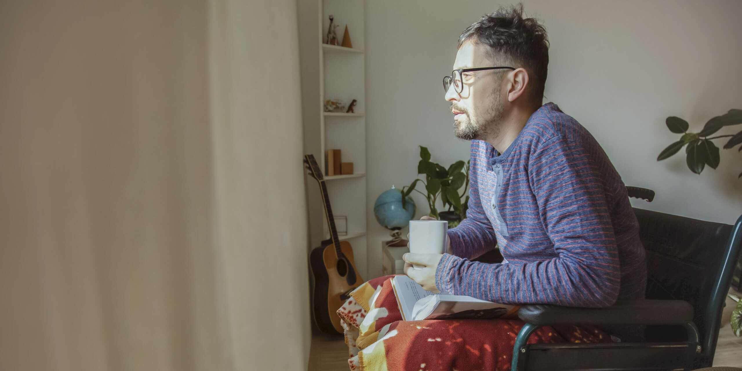 A young white man sits in a wheelchair in front of a window, looking out while holding a coffee mug and open book on his lap.