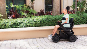 A young Black woman sits in a wheelchair outside on a cobblestone pathway in front of bushes and trees. She is wearing a green striped dress and white sneakers.