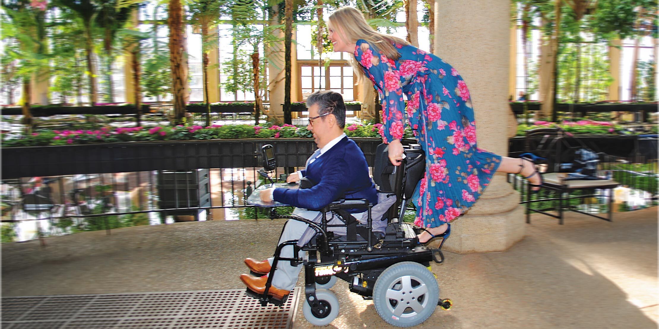 Santino sits in his wheelchair while Gill stands on the back with her foot kicked back behind. They are in a public garden and Santino is wearing a blue suit and Gill is wearing a floral blue dress.