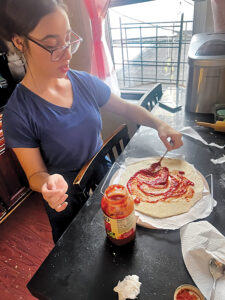 Leah Zelaya stands at a table while spreading pizza sauce on rolled out dough.