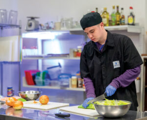 A young man chops vegetables in a professional kitchen. He is wearing a black uniform, black hat, purple long-sleeve undershirt and blue plastic gloves.