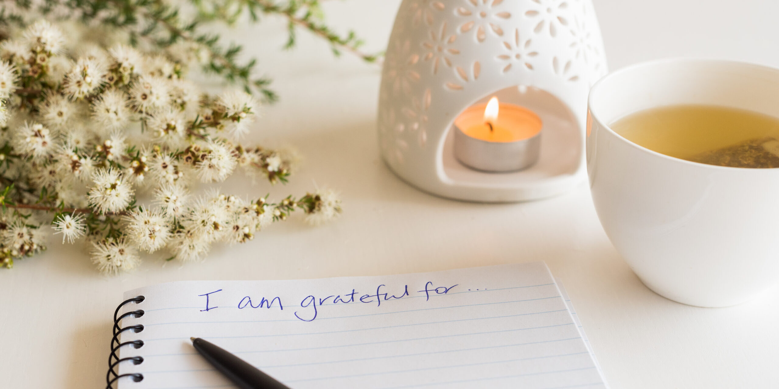 Close up of handwritten text "I am grateful for..." in foreground with notebook, pen, cup of tea, flowers and oil burner in soft focus