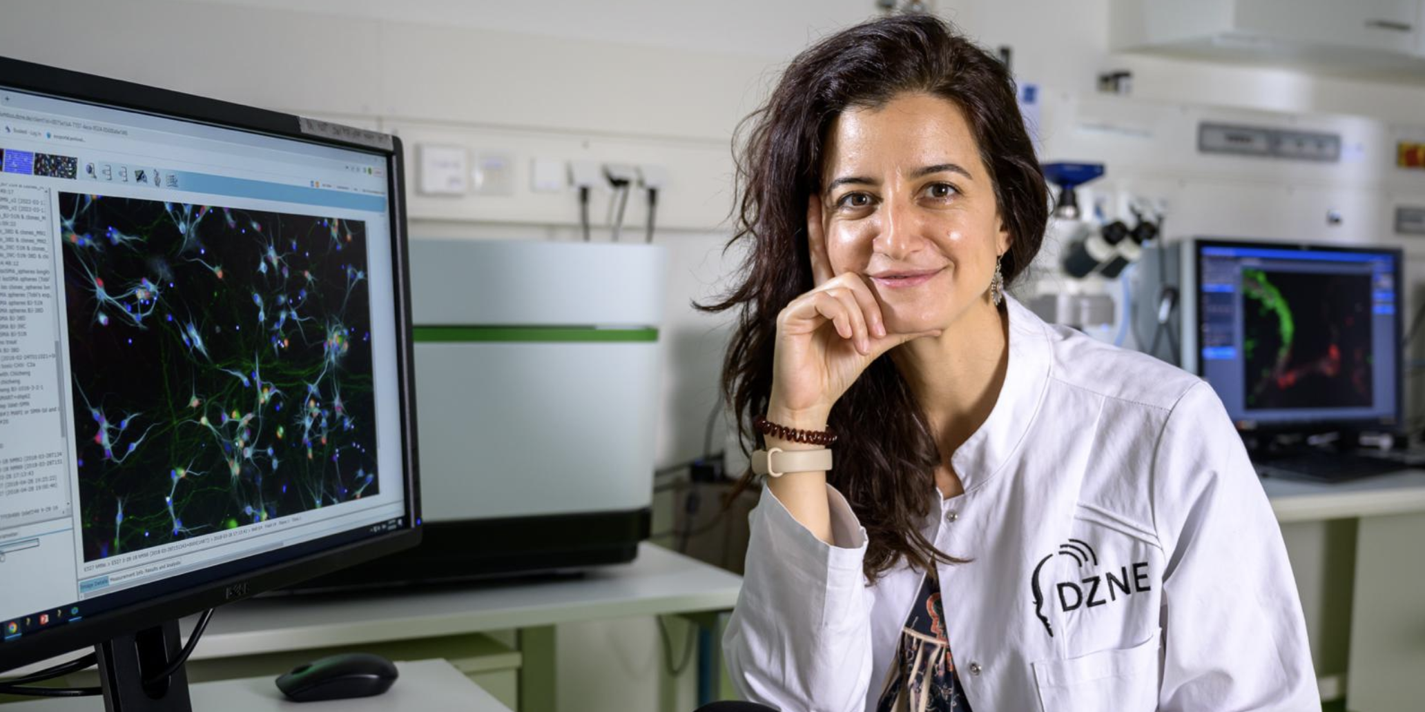 A researcher in a white lab coat with long brown hair rests her chin on her hand next to a computer with science research on the screen