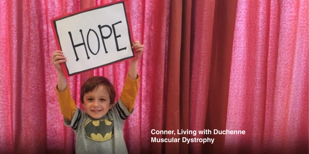 A young boy in a batman t shirt holds up a sign that says HOPE in front of a red curtain