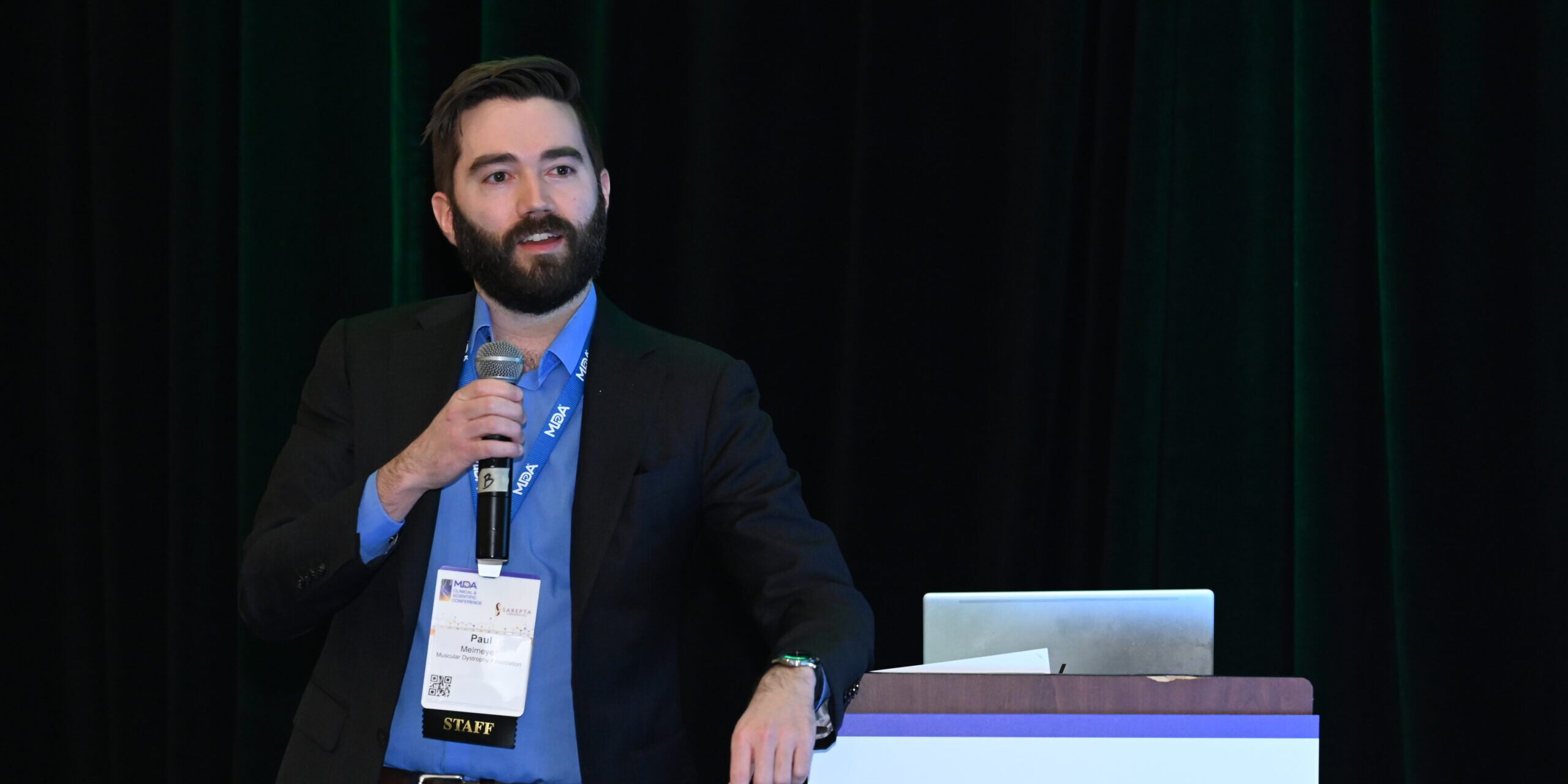 A man with a brown hair and a brown beard holds a microphone on stage at a conference