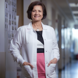 A scientist with dark brown shoulder length hair wearing a white lab coat over a pink, black, and white dress smiles with her hands in her pocket