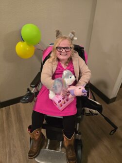 A young girl wearing glasses, black pants, and a pink dress and seated in a wheelchair with balloons attached smiles while holding two stuffed animals