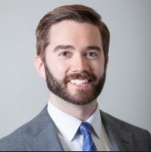 Headshot of a white man with brown hair and a brown beard, wearing a white collared shirt, blue tie, and gray jacket