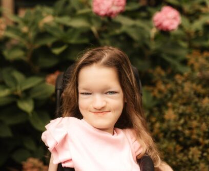 A young woman with brown hair smiles in front of a rose bush