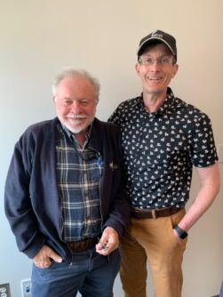 A man in a plaid shirt and a blue jacket with white hair and a white beard stands next to another man in a patterned short sleeve collared shirt, baseball hat, and glasses