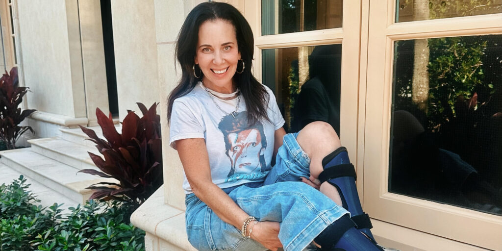 Estela Luna, a woman with light skin and long dark hair, sits outdoors on a stone bench with her feet propped up, revealing blue and black AFO braces from below her knees to her ankles. She wears silver sneakers, light blue jeans, and a David Bowie T-shirt, appearing relaxed in the setting.