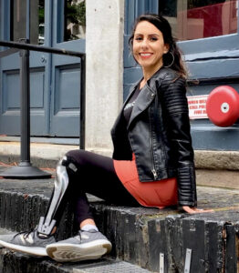 Sitting on black-painted stairs in an outdoor urban environment, Estela demonstrates how AFOs can be stylish. She’s wearing a black leather jacket over a black T-shirt, a red mini skirt over black ankle-length leggings, black AFOs over her leggings, and gray sneakers.