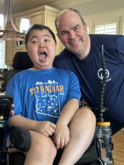 A young boy in a blue shirt sitting in a wheelchair makes a funny face while a man stands next to him smiling